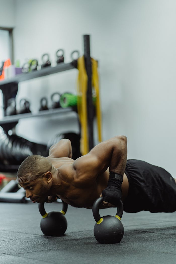 Muscular man performs push-ups using kettlebells in a gym setting, showcasing strength and fitness.